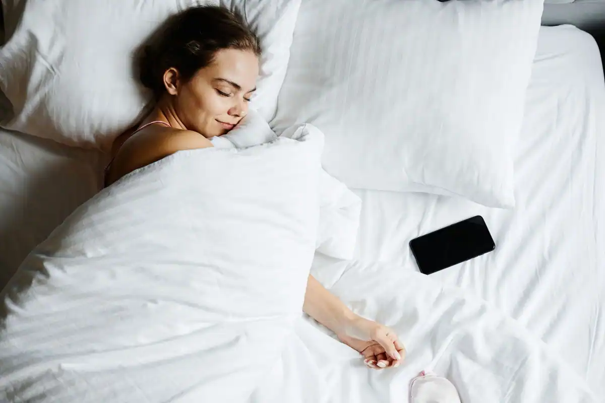 Tânără care doarme liniștit în pat, între cearșafuri albe, cu telefonul alături