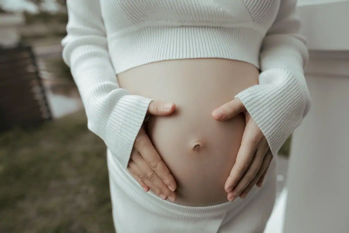 Femeie însărcinată care își ține burta cu ambele mâini, într-o ținută albă