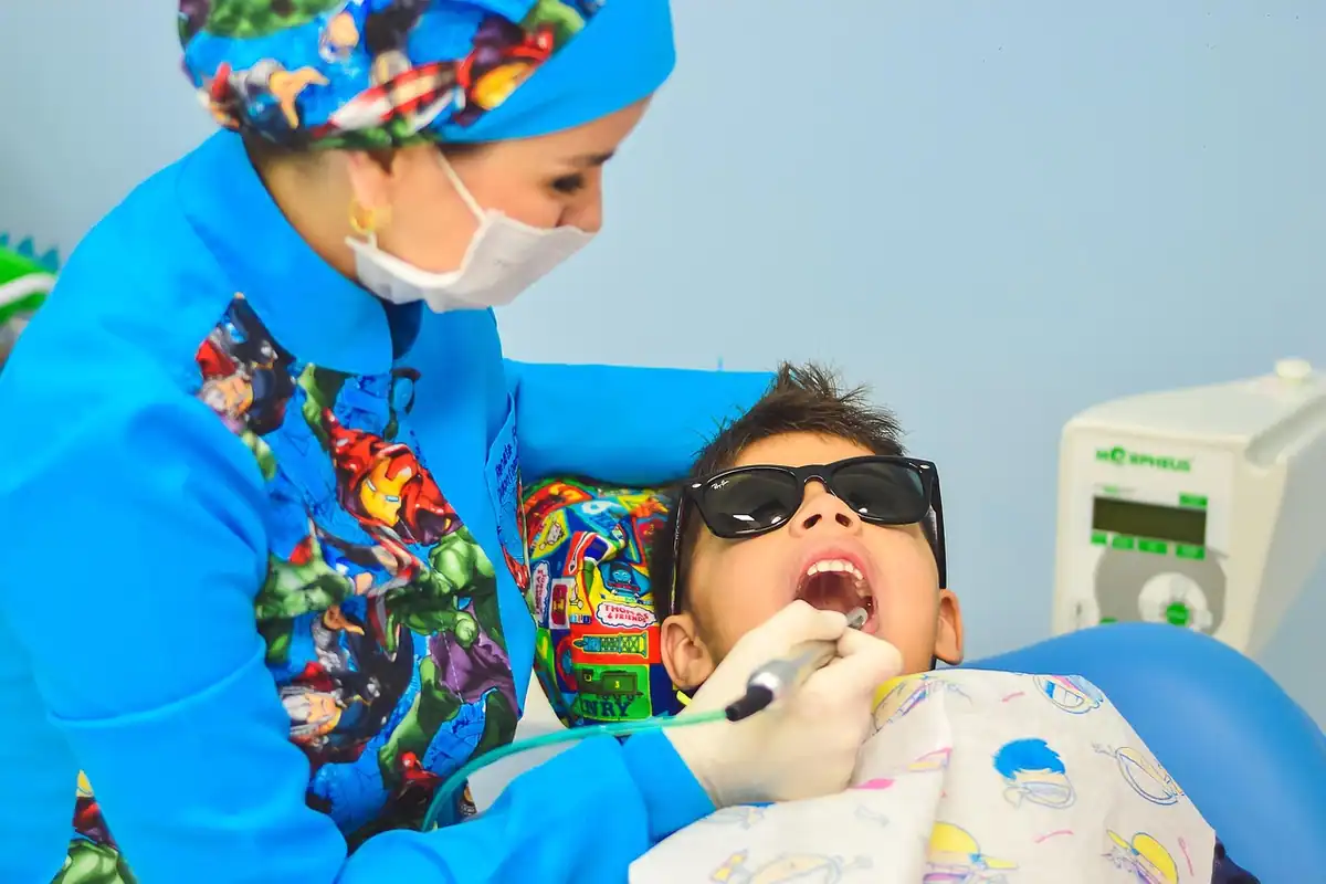 Medic stomatolog cu mască și halat colorat care examinează un băiețel cu ochelari de protecție